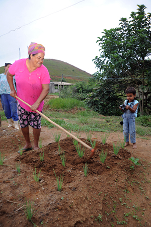 Senhora cava um chão de terra com plantações. Do lado direito dela, uma criança pequena com uma máquina de fotografar em mãos