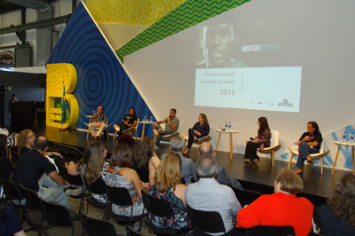 Seis pessoas, entre mulheres e homens, estão sentados em cadeiras na cor branca, em cima de um palco preto. À frente deles, uma plateia presta atenção ao que está sendo discutido. Atrás, um painel nas cores do Brasil, com a projeção de um slide e ao lado do palco uma letra 'B' grande na cor amarela.