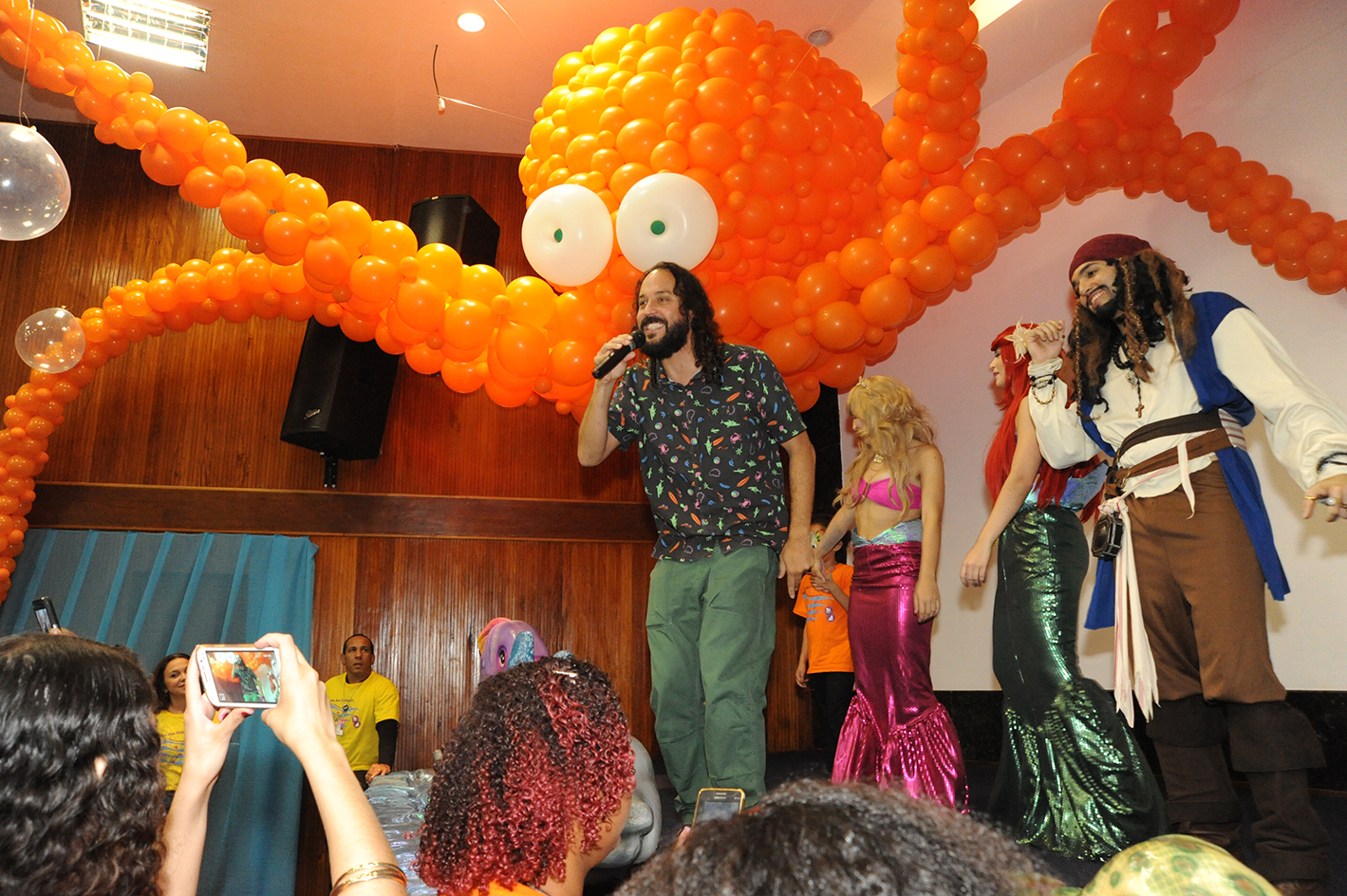 Gabriel, o Pensador está em cima do palco com camisa com desenho florido e calça na cor verde. Atrás dele, está um rapaz vestido como o personagem Jack Sparrow, um pirata. Ao lado há duas mulheres: uma vestida como a princesa sereia Ariel, com longo cabelo vermelho, blusa verde e saia longa verde imitando uma cauda de peixe. A outra mulher, com longos cabelos loiros, veste roupa temática na cor rosa. Em cima deles, figura um polvo gigante feito de bolas de festa na cor laranja