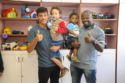 Os representantes do time distribuíram autógrafos e interagiram com pacientes Um homem do lado esquerdo de blusa cinza e calça preta segura uma criança de cabelos enrolados, de blusa colorida e bermuda jeans. Do lado direito, um outro homem também de blusa cinza e calça jeans segura uma bebê que veste uma blusa azul e branca e uma bermuda jeans.