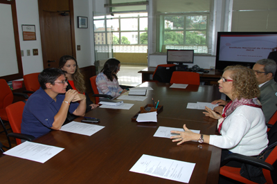 Durante conversa, Carina Vance abordou o sistema de saúde do Equador Cinco pessoas estão sentadas em volta de uma mesa marrom, entre mulheres e homens. Em frente a eles, descansam papéis sobre a mesa e canetas. Os presentes estão conversando e logo do lado da mesa existe uma TV com a foto de um slide.